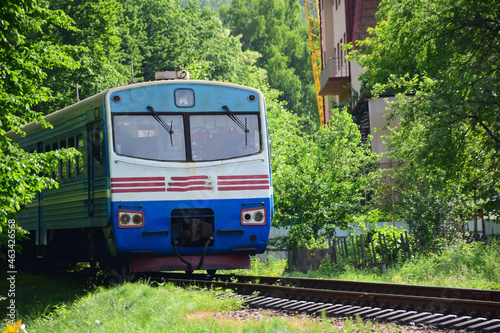The electric train leaves from afar from behind the trees to the railway station. Only the locomotive is visible