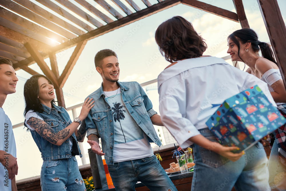 We have something special for you. Young woman is holding a gift box for her friend behind her back while standing on the roof with her friends We have something special for you. Young woman is holding a gift box for her friend behind her back while standing on the roof with her friends