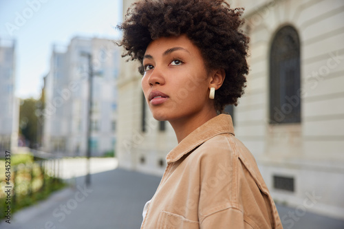 Sideways shot of thoughtful beautiful young woman with curly bushy hair looks into distance has charming look dressed in beige jacket strolls outdoors against blurred background blank copy space photo