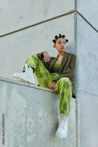 Vertical shot of hipster girl with bright makeup dressed in green fashionable clothes white boots poses against grey wall looks away thoughtfully spends free time in urban place. Youth lilfestyle