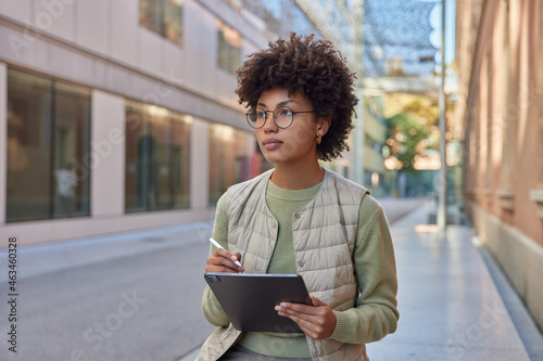 Creative beautiful woman with curly hair holds digital tablet draws sketches with stylus look away pensively wears round spectacles casual jumper and vest creats illustration on digital device photo