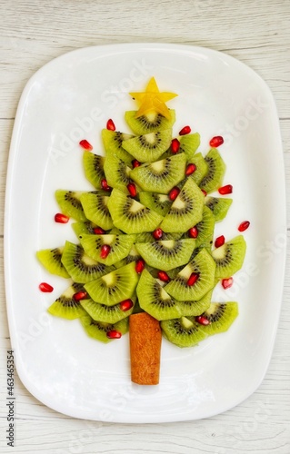 Fruit Christmas tree made it from kiwi fruit and pomegranate on plate with white wood background.Healthy vegan art food idea for Christmas party.Top view.Copy space