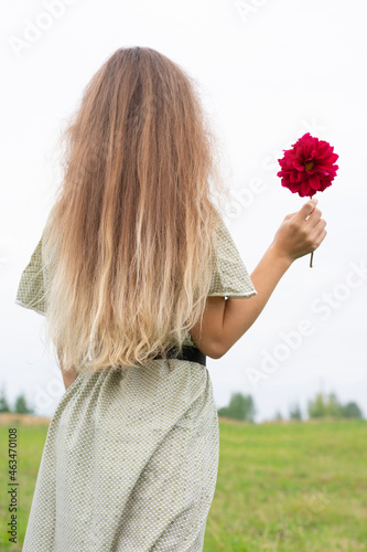Young blonde long haired woman with burgundy colored abloom dahlia flower in hand standing back and looking far away on horizon. Concept and metaphor of loneliness, love and hope