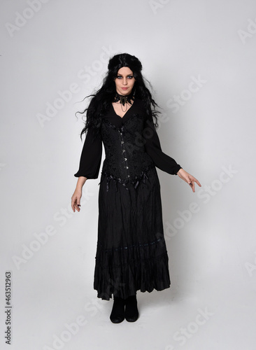 Full length portrait of dark haired woman wearing black victorian witch costume with corset, standing pose with gestural hand movements, against studio background.