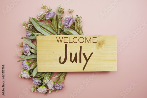 Welcome July text on wooden board with flowers frame on pink background