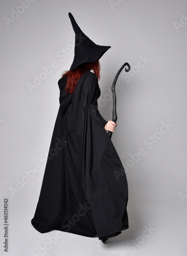 Full length portrait of dark haired woman wearing black victorian witch costume with cloak and pointy hat. standing pose, back view, with gestural hand movements, against studio background.