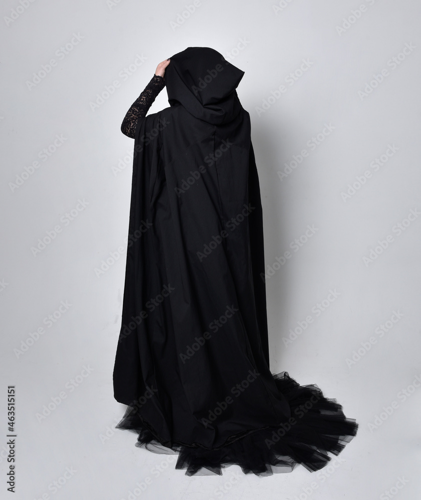 Full length portrait of dark haired woman wearing  black victorian witch costume with  cloak and pointy hat.  standing pose, back view,  with  gestural hand movements,  against studio background.