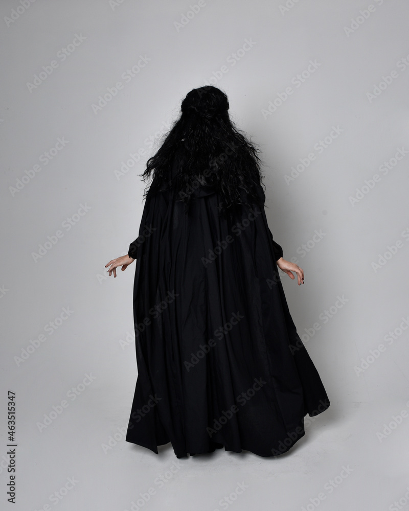 Full length portrait of dark haired woman wearing  black victorian witch costume with  cloak and pointy hat.  standing pose, back view,  with  gestural hand movements,  against studio background.