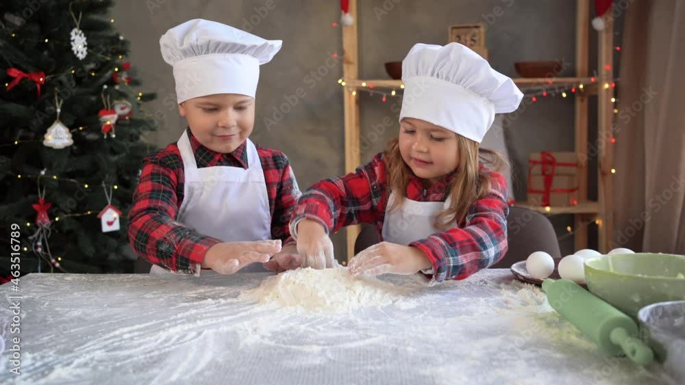 Funny kids in the kitchen prepare raw flour dough for baking Christmas gingerbread cookies. Brother and sister play with flour dressed in the uniform Funny kids in the kitchen prepare raw flour dough for baking Christmas gingerbread cookies. Brother and sister play with flour dressed in the uniform
