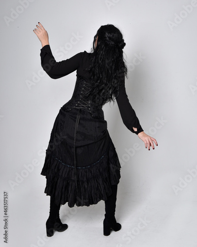 Full length portrait of dark haired woman wearing black victorian witch costume. standing pose, back view, with gestural hand movements, against studio background.