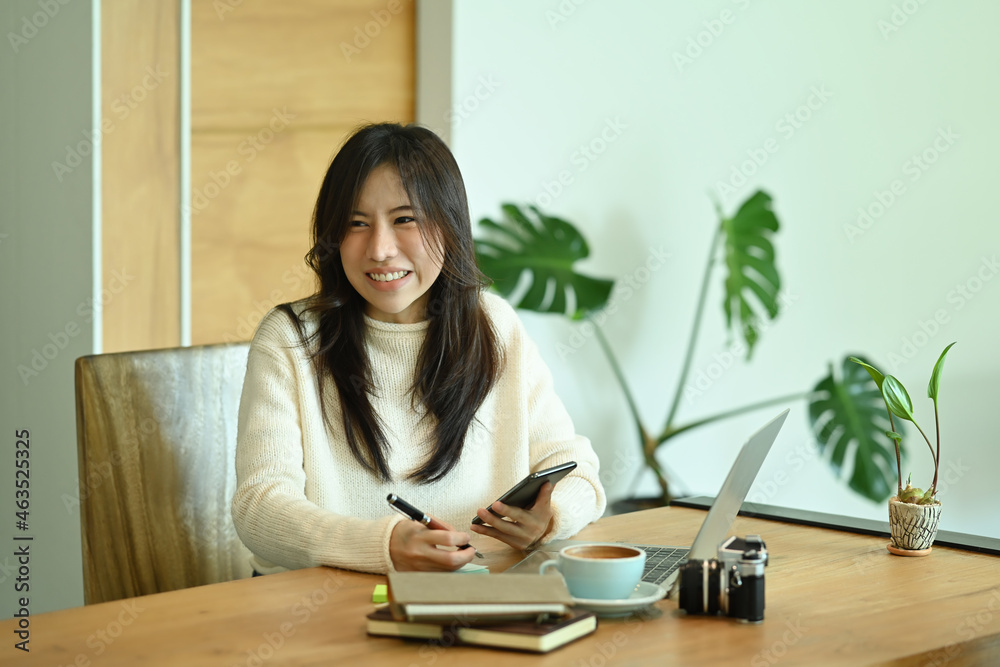Cheerful young woman holding mobile phone and working with computer laptop in office. Cheerful young woman holding mobile phone and working with computer laptop in office.
