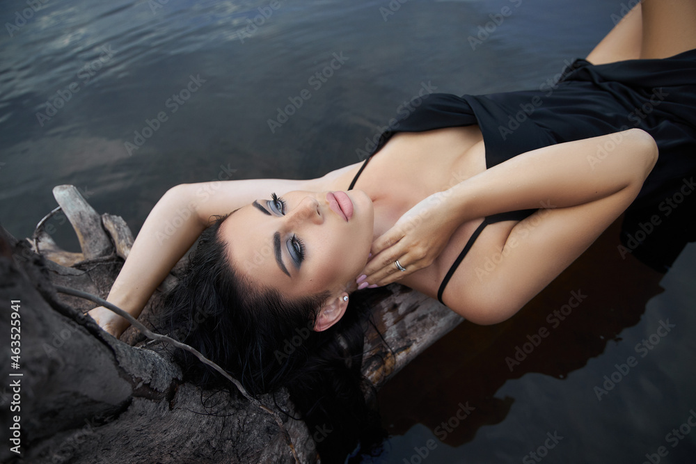 Fototapeta premium Beauty brunette woman in a black dress poses in a sea lake against a blue sky background. Long hair sexy woman and beautiful beauty makeup on her face