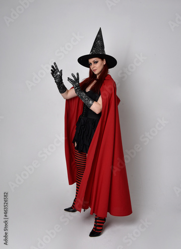 Full length portrait of dark haired woman wearing black victorian witch costume with a flowing cloak. standing pose, with gestural hand movements, against studio background.