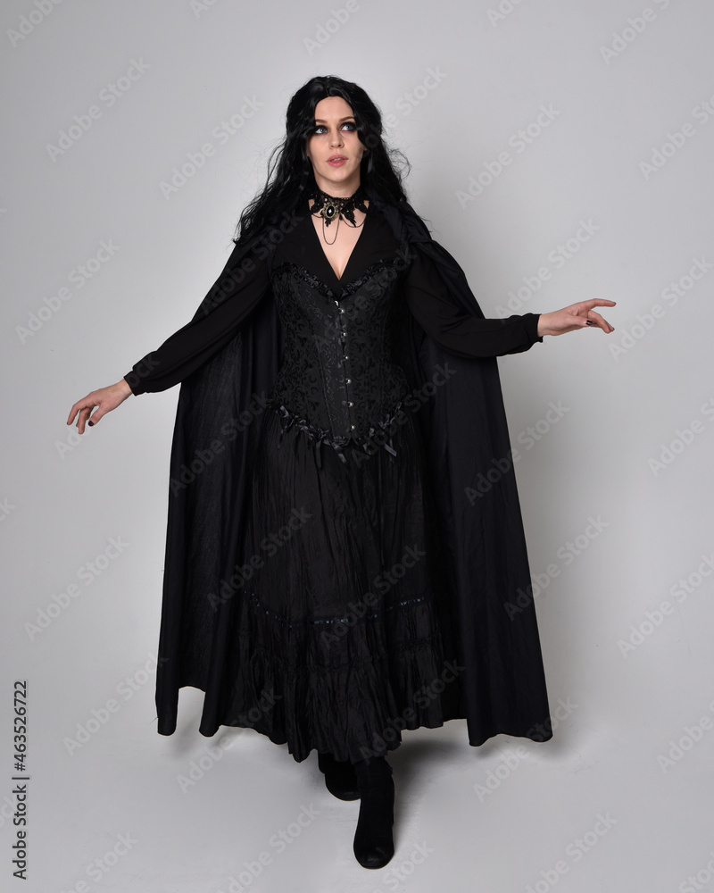 Full length portrait of dark haired woman wearing black victorian witch costume with a flowing cloak. standing pose, with gestural hand movements, against studio background. Full length portrait of dark haired woman wearing black victorian witch costume with a flowing cloak. standing pose, with gestural hand movements, against studio background.