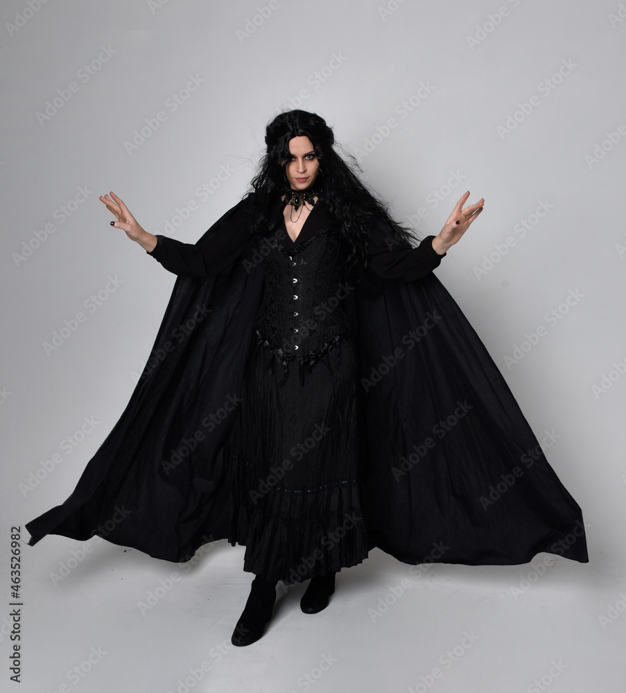 Full length portrait of dark haired woman wearing black victorian witch costume with a flowing cloak. standing pose, with gestural hand movements, against studio background. Full length portrait of dark haired woman wearing black victorian witch costume with a flowing cloak. standing pose, with gestural hand movements, against studio background.