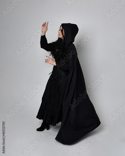 Full length portrait of dark haired woman wearing black victorian witch costume with a flowing cloak. standing pose, with gestural hand movements, against studio background.