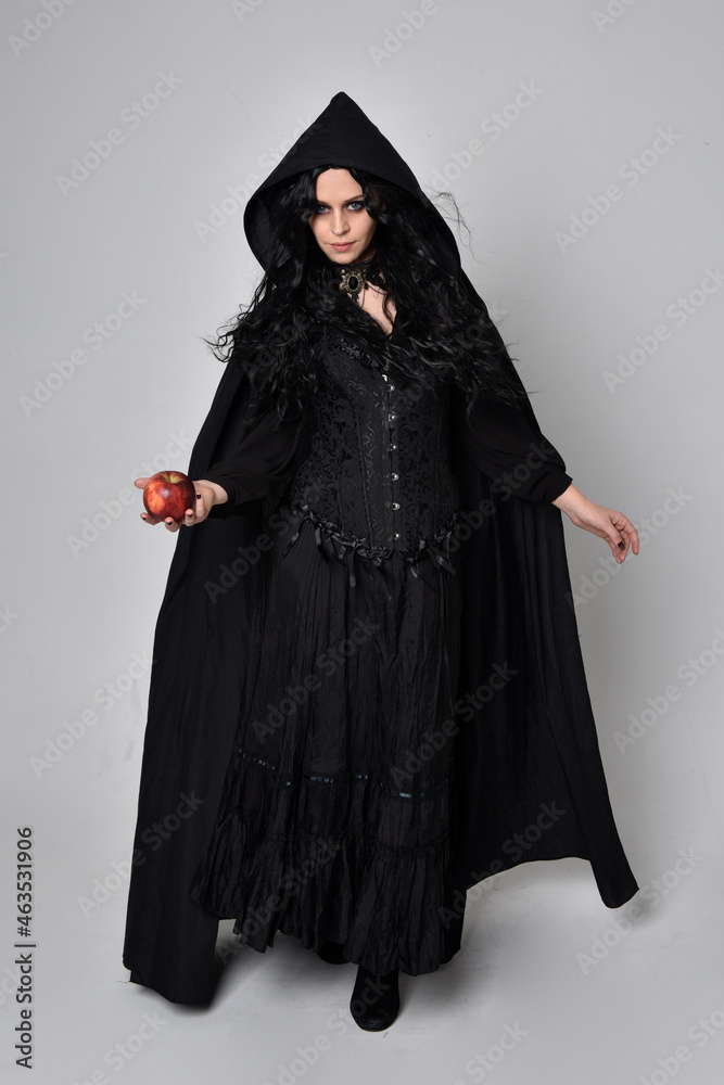 Full length portrait of dark haired woman wearing  black victorian witch costume  standing pose, with  gestural hand movements,  against studio background.