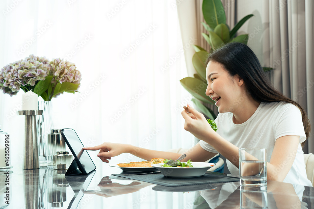 Asian young lady picks up her favorite series on her tablet while happily scooping a salad into her mouth at home.