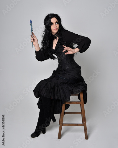 Full length portrait of dark haired woman wearing black victorian witch costume. sitting pose on a chair, with gestural hand movements, against studio background.