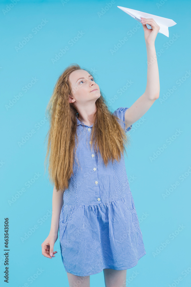 Travel Concepts. Caucasian Teenage Girl in Blue Checked Dress Launching Origami Paper Plane Over Blue Background.