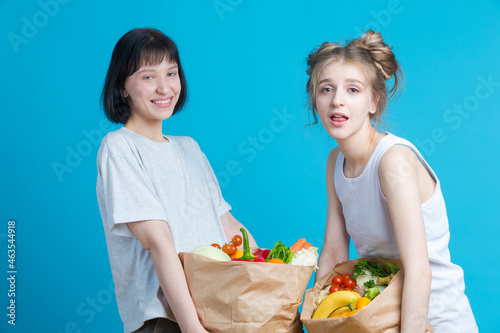 Two Positive Laughing Caucasian Girls Posing With Eco Paper Bags Filled With Grocery And Vegetables Over Blue Background