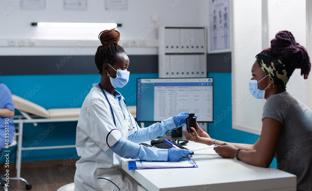 Obraz premium African american doctor with protective face mask against covid19 explaining medication treatment to sick patient holding pills bottle during clinical examination. Physician working in hospital office