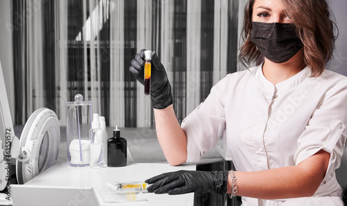 Schilderij op canvas Cosmetologist wearing black gloves, face mask and white uniform, sitting at the table holding blood tube with separated human plasma
