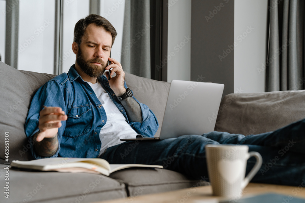 Fototapeta premium Bearded man talking on cellphone and using laptop while working at home