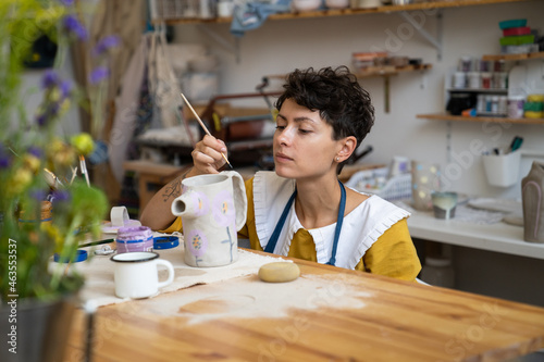 Creative process in pottery studio: young craftswoman or self employed ceramist decorating potter jug for sale in handmade crockery store with paint, hand drawn ornament. Art hobby and leisure concept photo