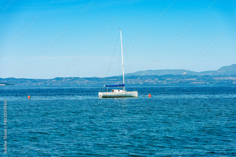 White sailing boat moored in front of the coast of Lake Garda (Lago di Garda), near the small village of Lazise, tourist resort in Verona province, Veneto, Italy, Europe. On horizon Lombardy coast. White sailing boat moored in front of the coast of Lake Garda (Lago di Garda), near the small village of Lazise, tourist resort in Verona province, Veneto, Italy, Europe. On horizon Lombardy coast.