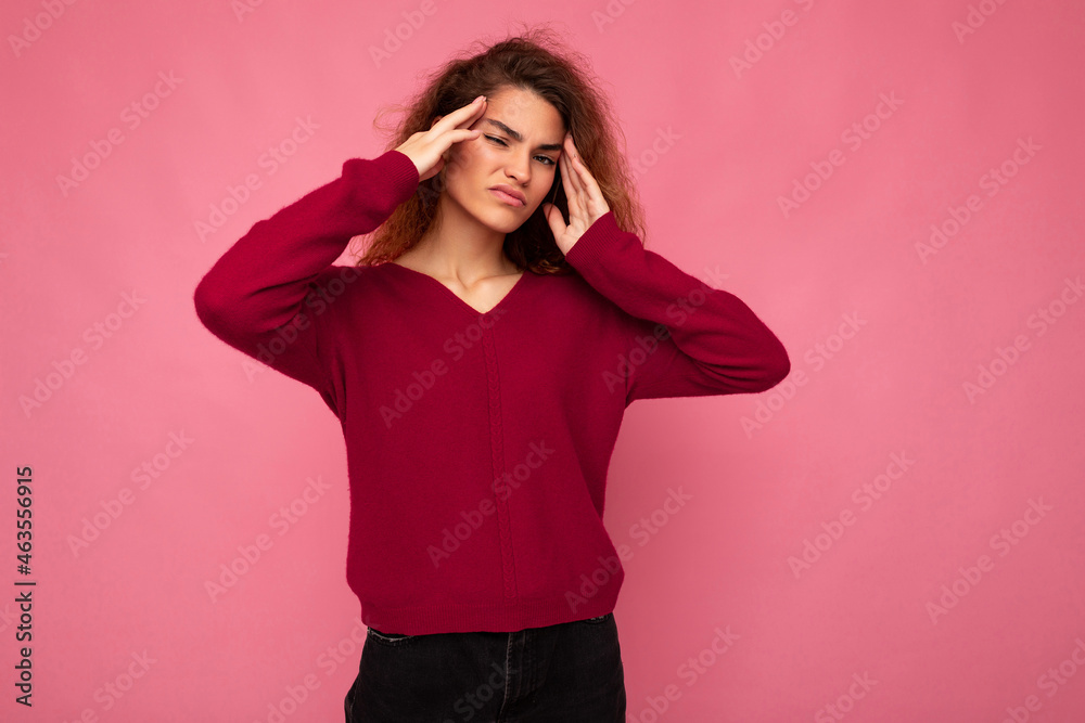 Young emotional attractive brunette curly woman with sincere emotions wearing trendy pink pullover isolated on pink background with copy space and having headache Young emotional attractive brunette curly woman with sincere emotions wearing trendy pink pullover isolated on pink background with copy space and having headache