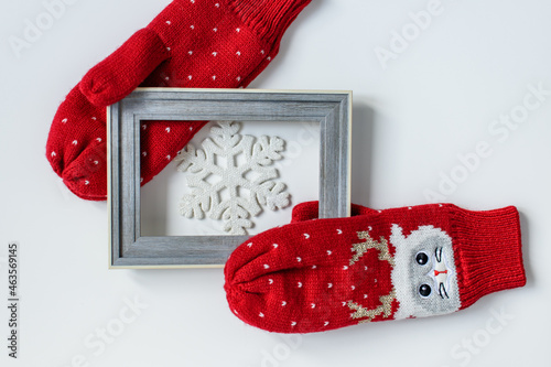 A pair of red woolen knitted soft mittens with cat faces and deer antlers holding a snowflake in a wooden frame on a white background. Christmas and New Years concept. Flat lay, top view.