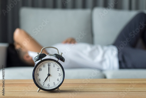 .The clock is placed on a wooden table with a young man in a white T-shirt lying on the sofa in the living room at home. He felt a headache and discomfort after working at home for a long time.