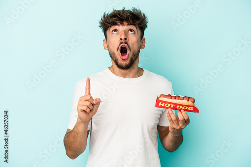 фотографія Young arab man holding a hotdog isolated on blue background pointing upside with opened mouth
