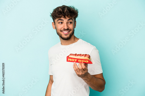 Принти на полотні Young arab man holding a hotdog isolated on blue background looks aside smiling, cheerful and pleasant