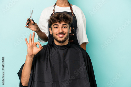 Young arab man ready to get a haircut isolated on blue background cheerful and confident showing ok gesture.