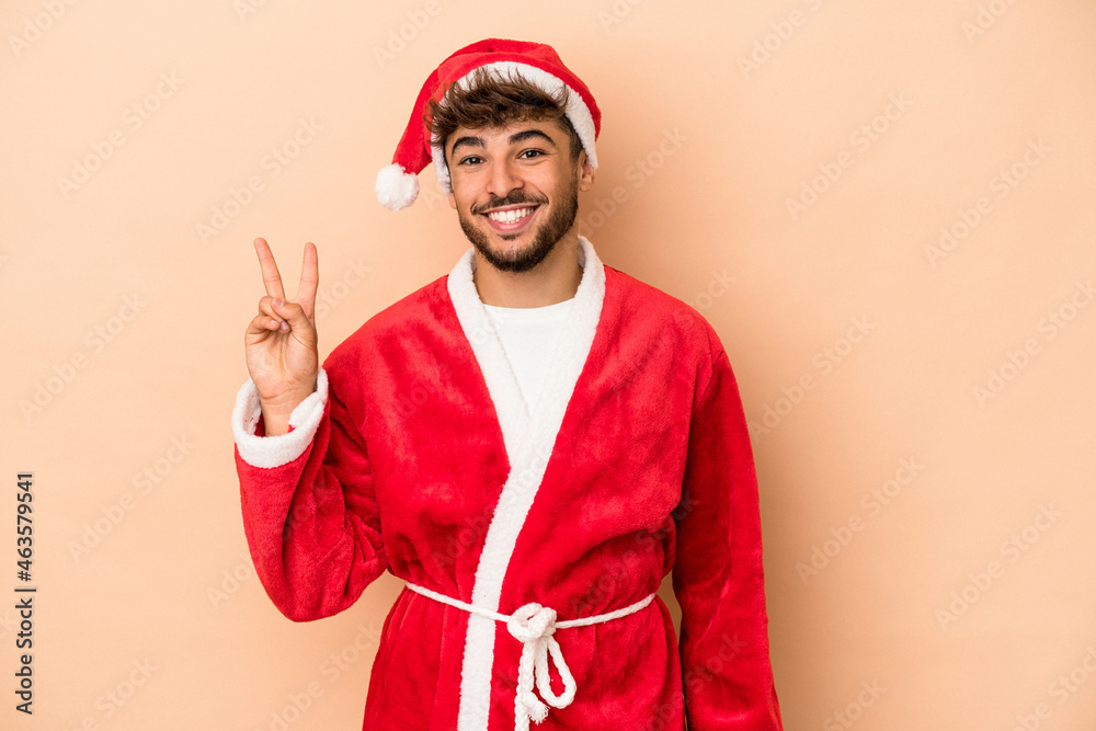 Young arab man disguised as santa claus isolated on beige background joyful and carefree showing a peace symbol with fingers. Young arab man disguised as santa claus isolated on beige background joyful and carefree showing a peace symbol with fingers.