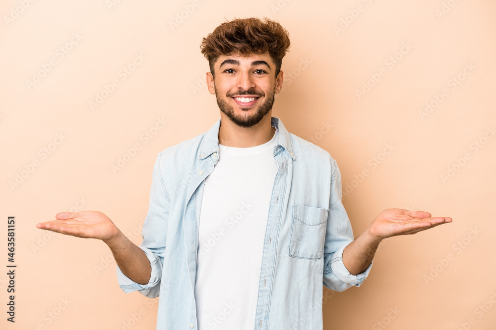 Young arab man isolated on beige background makes scale with arms, feels happy and confident.