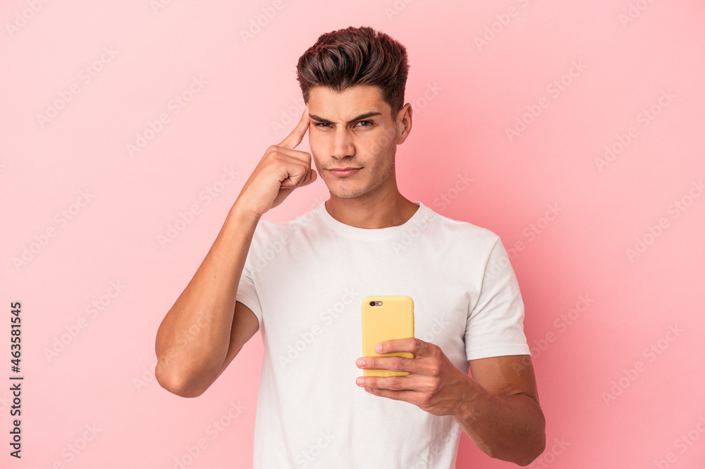 Young caucasian man holding a mobile phone isolated on pink background pointing temple with finger, thinking, focused on a task. Young caucasian man holding a mobile phone isolated on pink background pointing temple with finger, thinking, focused on a task.