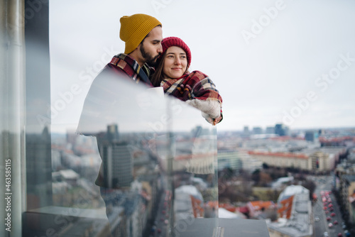 Young couple wrapped in blanket standing and hugging outdoors on balcony, shot through glass. © Halfpoint