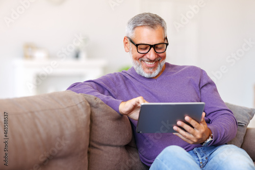 Handsome happy senior man using digital tablet while relaxing resting on sofa at home