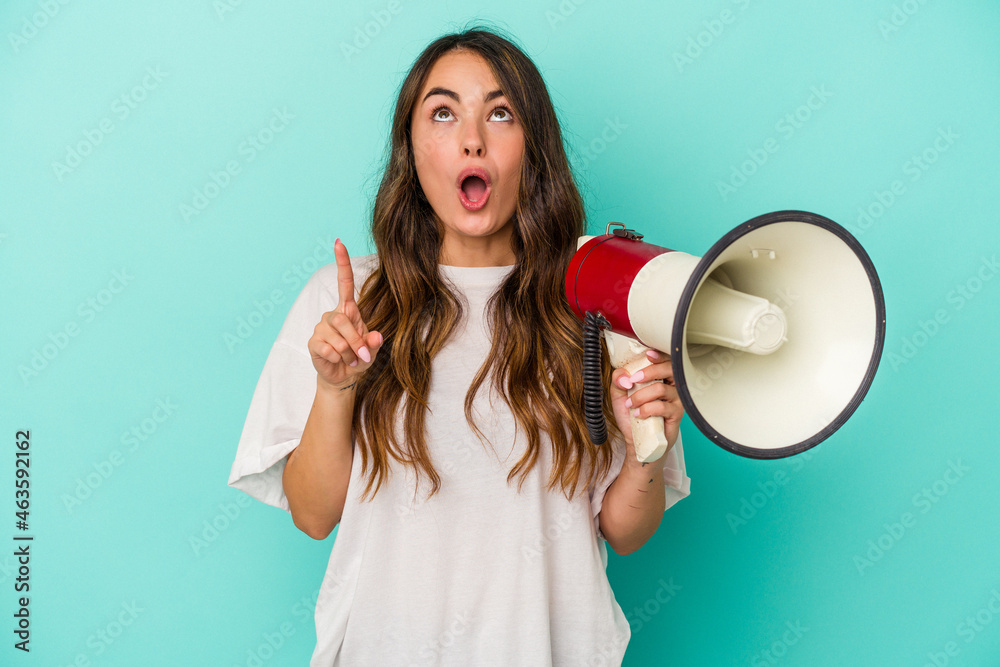 Young caucasian woman holding a megaphone isolated on blue background pointing upside with opened mouth. Young caucasian woman holding a megaphone isolated on blue background pointing upside with opened mouth.