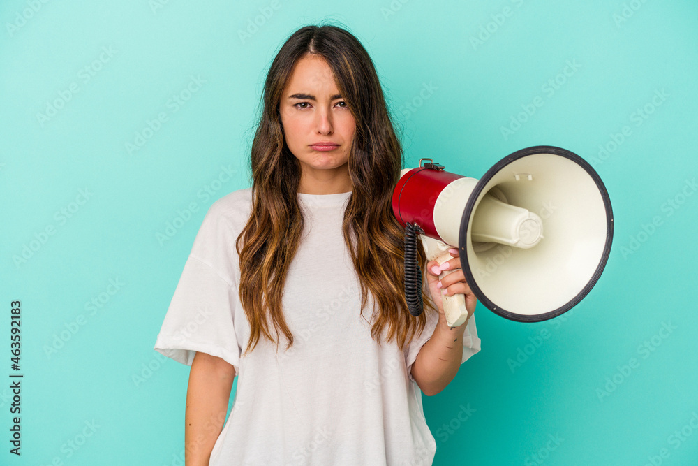 Young caucasian woman holding a megaphone isolated on blue background shrugs shoulders and open eyes confused. Young caucasian woman holding a megaphone isolated on blue background shrugs shoulders and open eyes confused.
