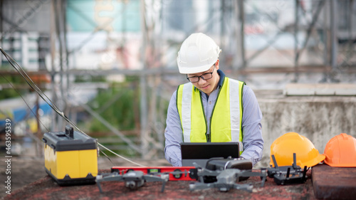 Asian engineer man working with drone, laptop and working tools at construction site. Male worker using unmanned aerial vehicle (UAV) for land and building site survey in civil engineering project.