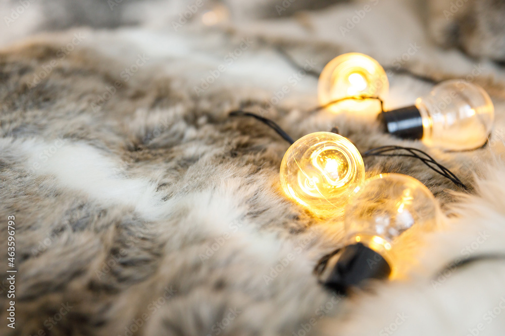 Fototapeta premium Garland lamps lie on a fluffy, cozy fur blanket. Coziness and atmospheric mood in the home interior. New Year and Christmas background. Place for text or product, copy space