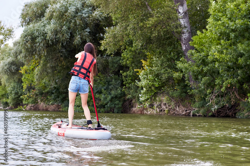 Caucasian woman rowing on stand up paddle board, SUP alone in river near green trees. Back view