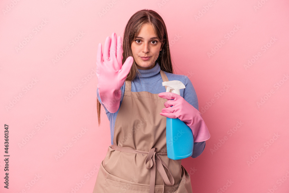 Young caucasian cleaner woman holding spray isolated on pink background standing with outstretched hand showing stop sign, preventing you. Young caucasian cleaner woman holding spray isolated on pink background standing with outstretched hand showing stop sign, preventing you.