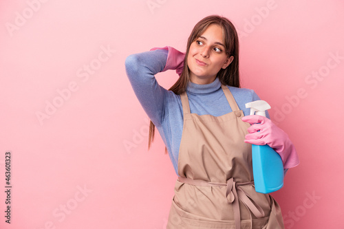 Young caucasian cleaner woman holding spray isolated on pink background touching back of head, thinking and making a choice.