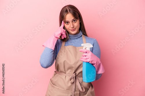 Young caucasian cleaner woman holding spray isolated on pink background pointing temple with finger, thinking, focused on a task.