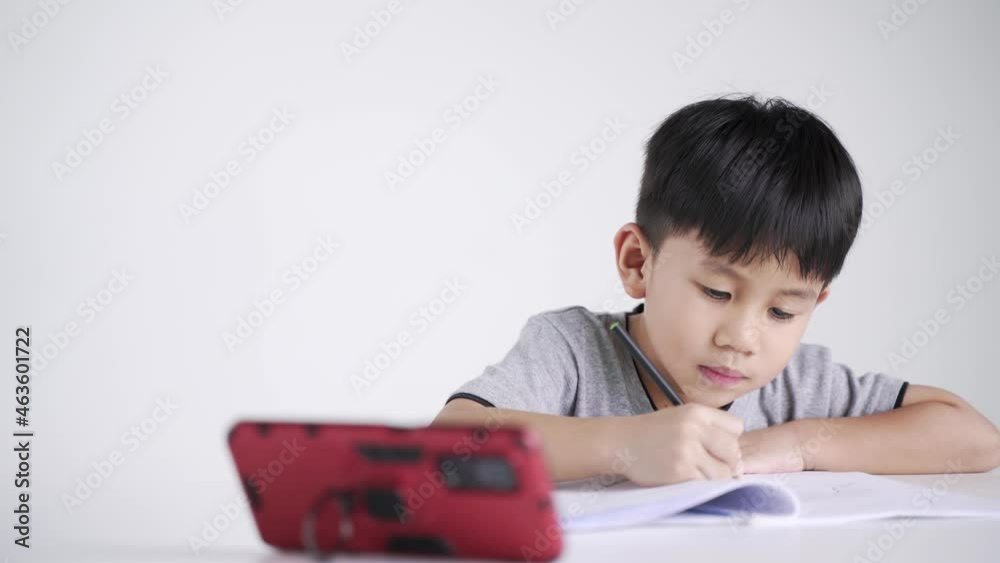 Asian boy about 5 years old writing while watching a mobile phone as studying online due to Covid 19 pandemic Asian boy about 5 years old writing while watching a mobile phone as studying online due to Covid 19 pandemic